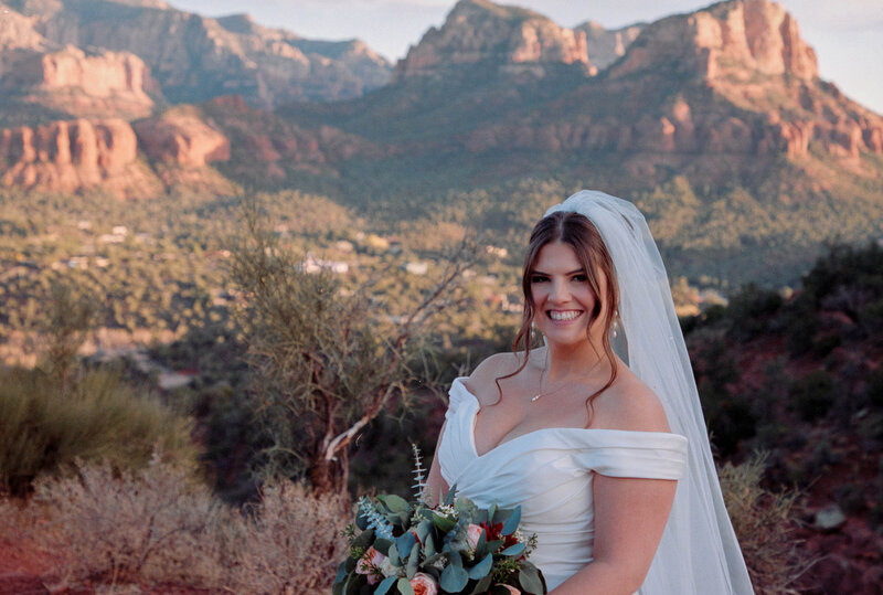 Bride in a veil with flower bouquet looking at the camera standing in front of Sedona red rock views