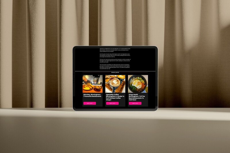 A tablet placed on a beige surface, displaying the B Town Social website. The screen shows the latest posts section, featuring articles about popular Birmingham eateries. Each post has an image of food and a title highlighting topics like favourite restaurants and speciality coffee shops. The background features beige curtains, creating a warm and inviting setting.