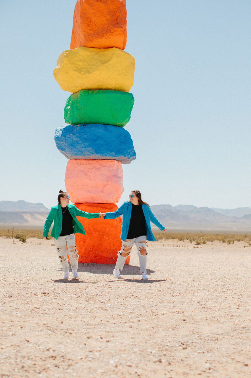 Two girls stand in front of rainbow rocks with blazers on while holding hands in the desert