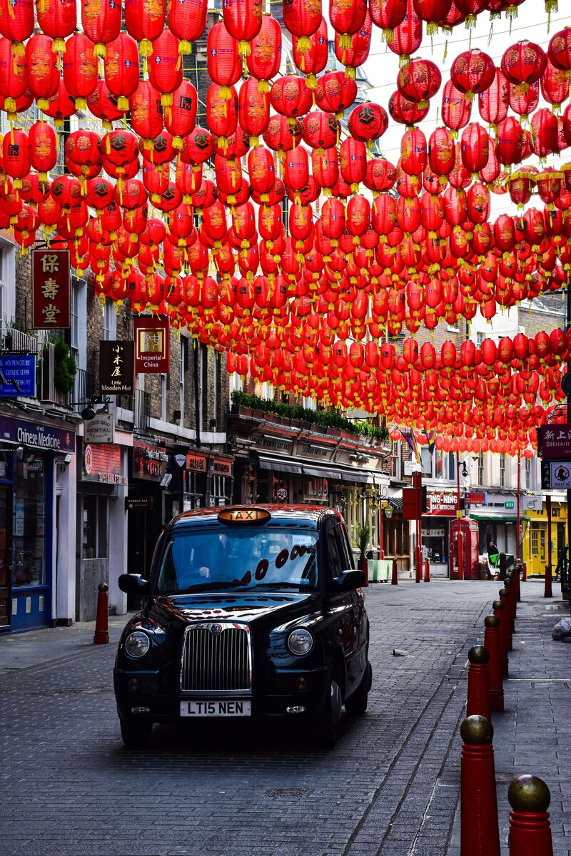 A bustling street in London's Chinatown, adorned with vibrant red lanterns strung overhead, creating a festive atmosphere. A classic black London taxi drives down the cobblestone road, showcasing the lively and diverse travel opportunities supported by vacation fundraising for charities.