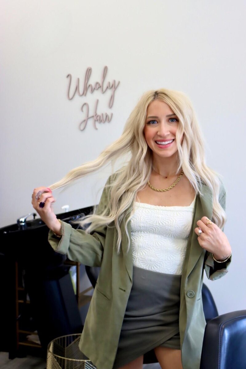 woman wearing white shirt and blazer with a skirt smiling while she touchese her hair standing in front of wholy hair sign in a salon suite