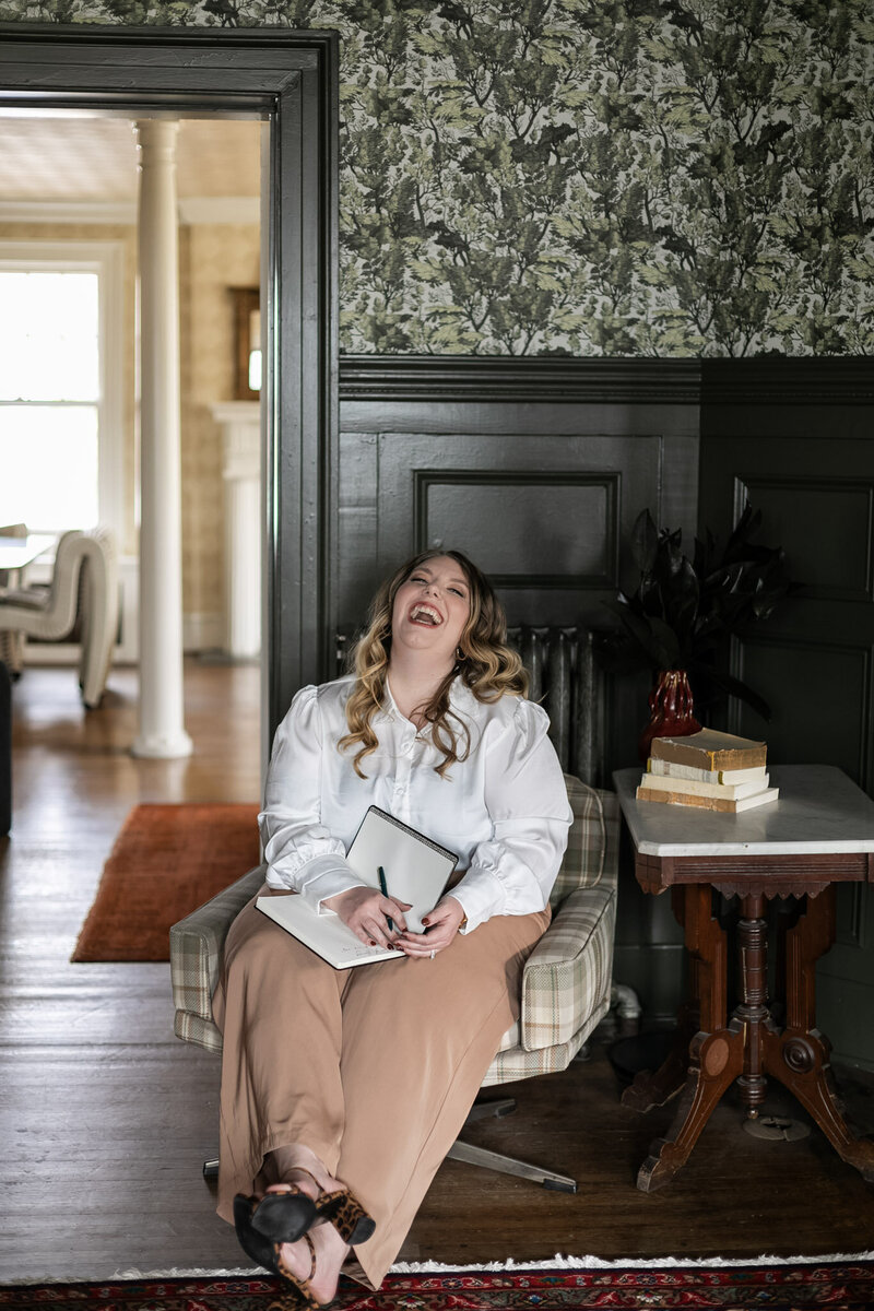 Portrait of Ashleigh Henry, high-achieving women’s coach, dressed in a white button down with khaki pants and cheetah print heels, representing the fullness of inhabited, integrated living while writing in her journal at The Blind Tiger.