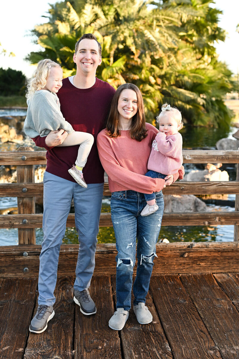 Parents standing on a wooden bridge each holding a small child on their hip.