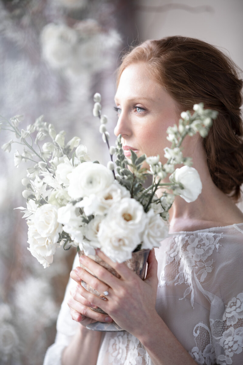 A bride in a lace dress stares off into the distance holding a wedding centerpiece with white flowers and branches