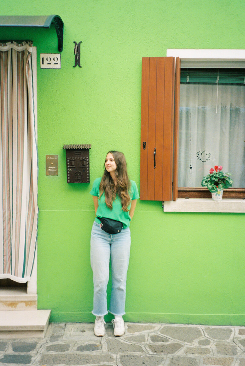 A portrait of Hannah, a Winnipeg wedding photographer, standing in front of a bright green wall, showcasing her creative personality.