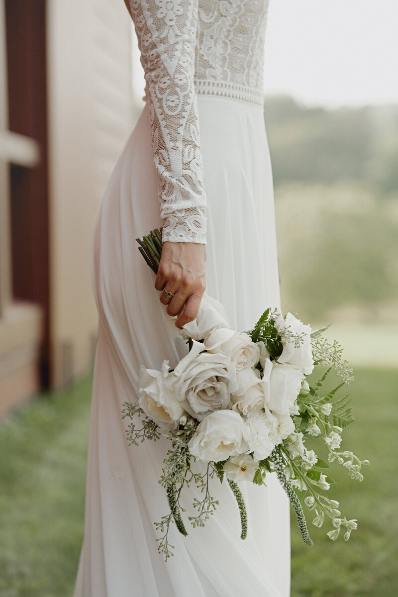 A bride in an an embroidered lace wedding gown standing outside a red barn holding a bridal bouquet of white and gray garden roses larkspur and veronica at her side