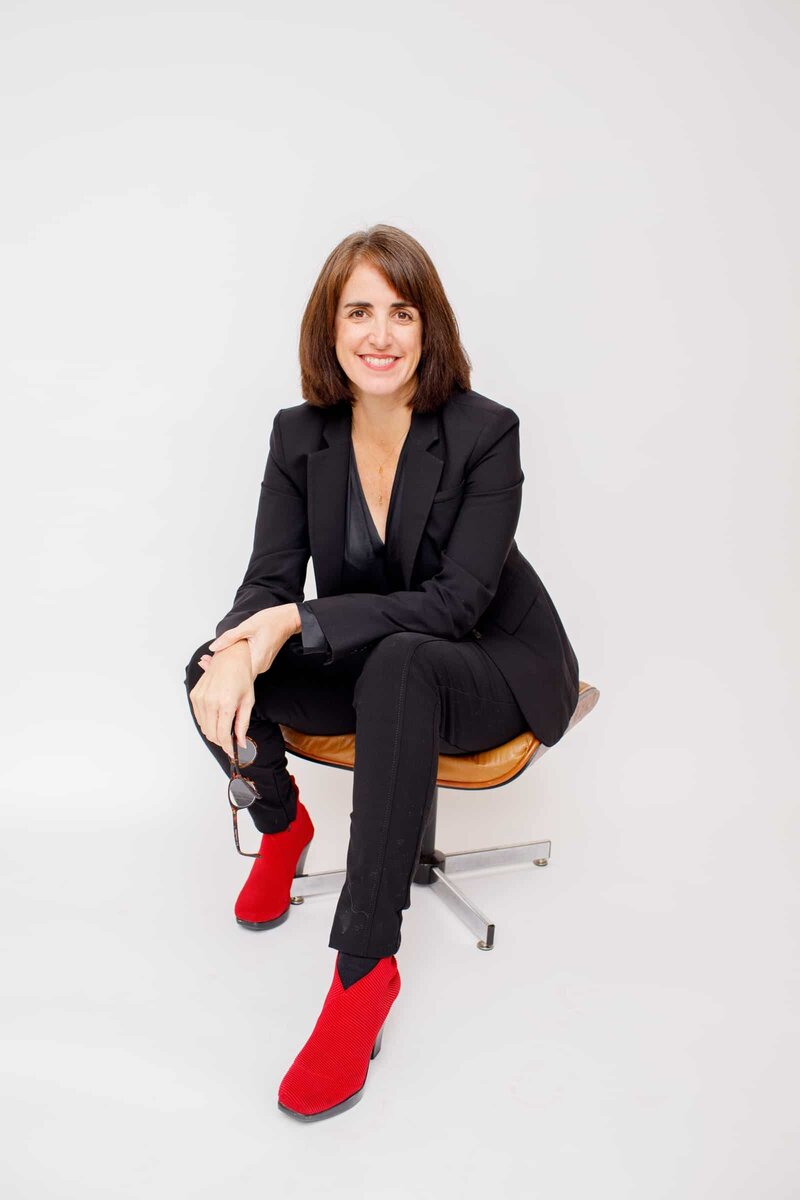 Woman wearing a black suit and red boots sitting in a white room