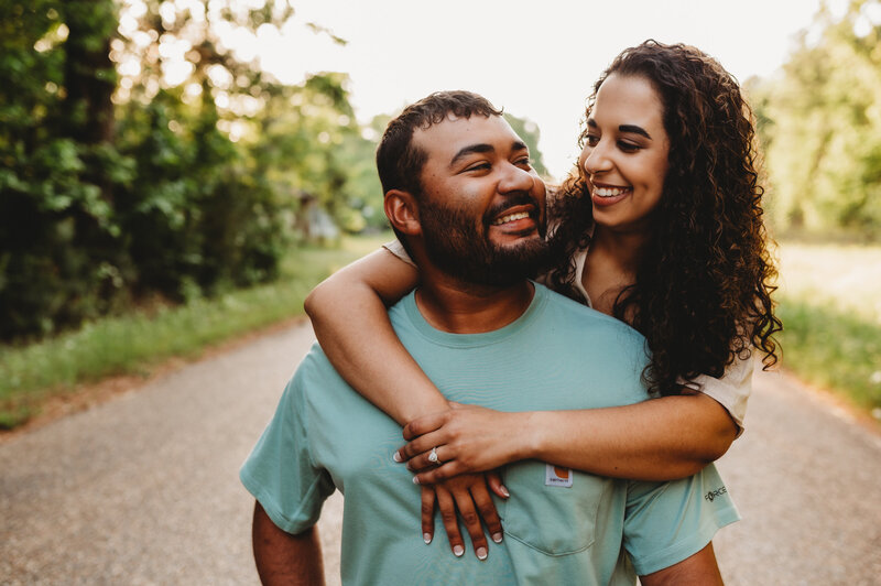 outdoor engagement session with man and woman on a path with woman piggy backing on man and looking over his shoulder and smiling down to him