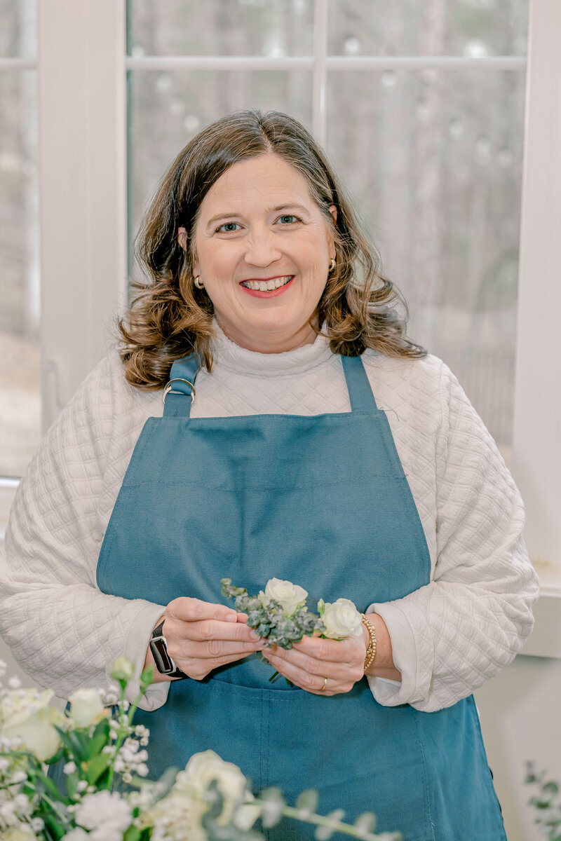 dark haired woman in apron smiling at camera holding white flowers