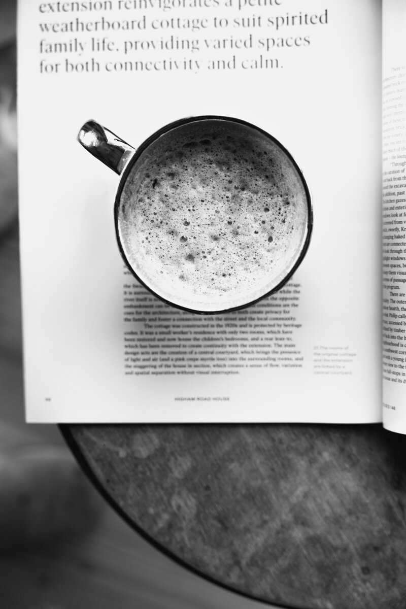 black and white image of frothy coffee in gold gilded cup on top of an open magazine with print on it on top of a wooden side table