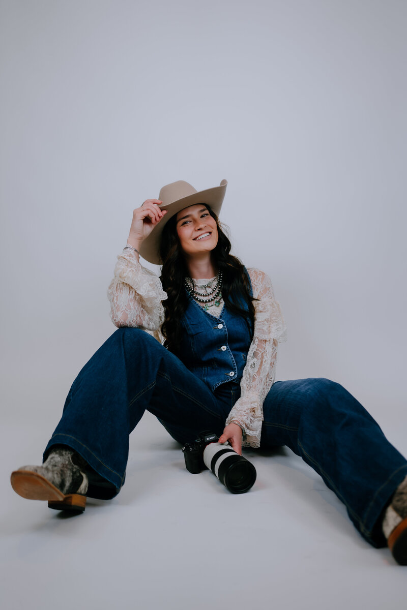 woman sitting on the ground with her camera while smiling and touching her cowgirl hat
