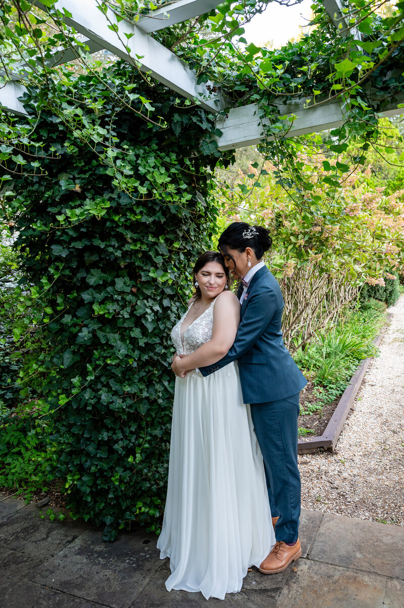 laotian bride in blue suit holding and nuzzling the hair of a curvy white bride in a spaghetti-strap gown with eyes closed and smiling