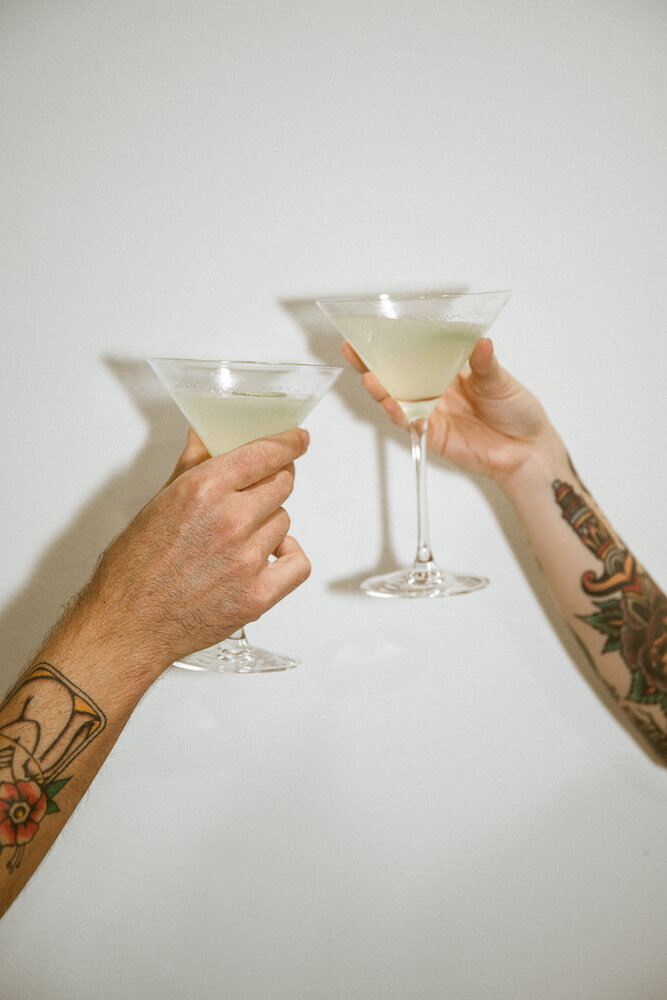 tattooed couple holding martini glasses against white wall