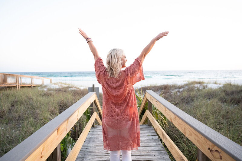 woman standing on dock in front of ocean with hands up in the air