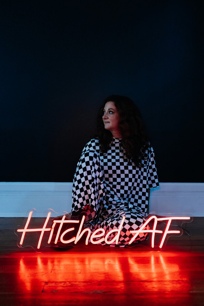 A woman sitting on the floor with a neon sign in front of them that reads "Hitched AF"