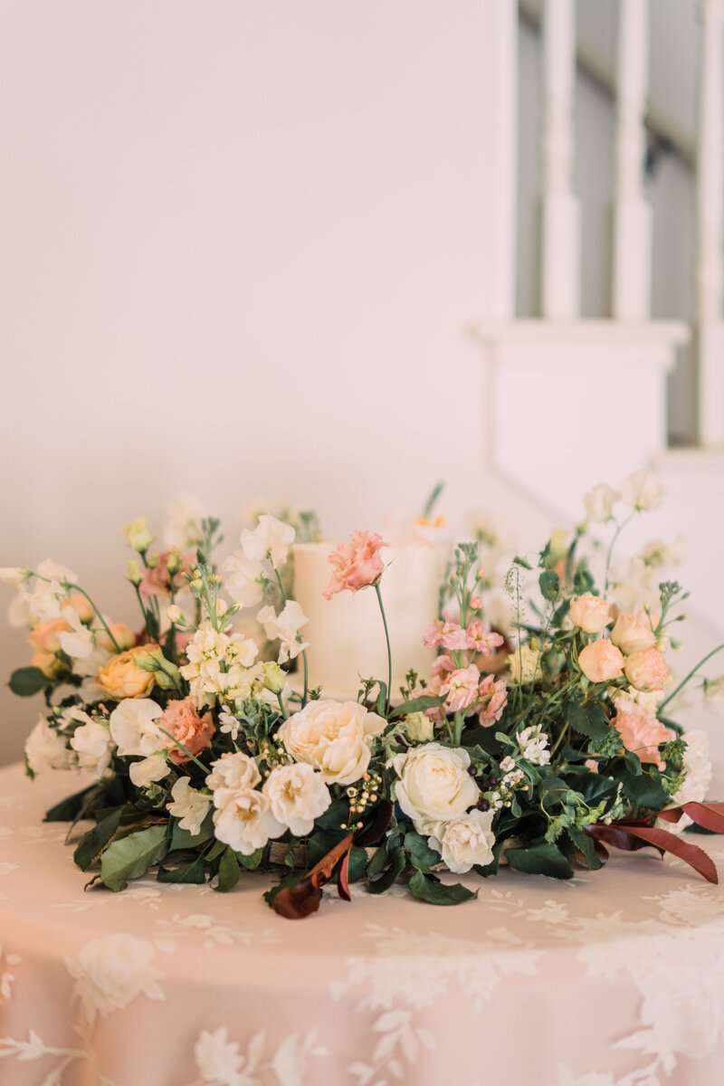 Cake surrounded by a garden wreath of roses & greenery sitting on pink linens with a lace overlay at Zingerman's Cornman Farms