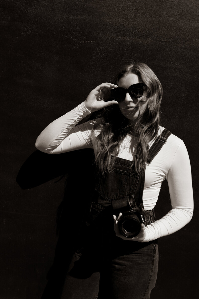 A black and white photo of a photographer while wearing sunglasses and holding her digital camera.