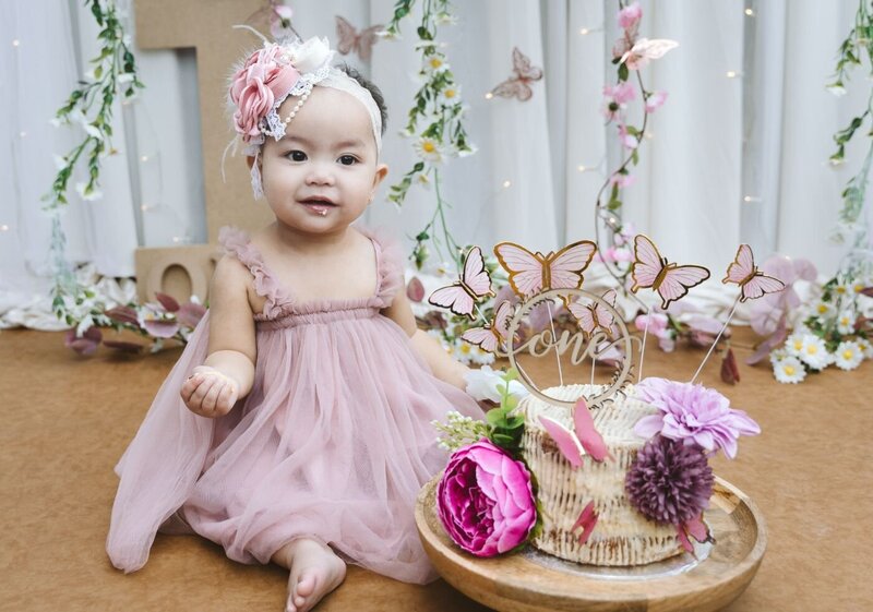 Baby girl sitting on a patterned pink blanket in a meadow, looking up at pastel balloons during an outdoor birthday shoot