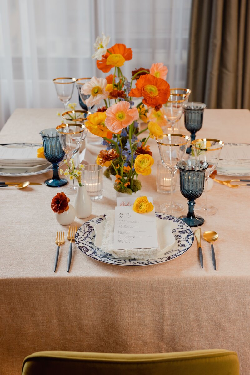 Luxurious wedding table setting with bright orange and yellow flowers, gold-rimmed glassware, and blue goblets on a soft linen tablecloth.