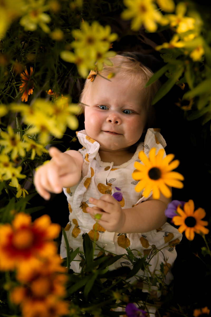 baby girl lays on a blanket surrounded by yellow, purple, and orange flowers pointing up at the camera