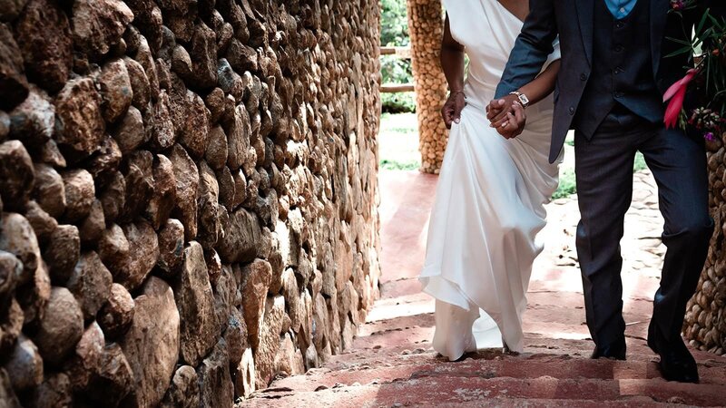 A bride and groom in wedding formalwear hold hands as the focal of the image while they walk up a set of red stairs with textured round pebble stone walls visible to the left of the frame and behind them.  They are off to meet the safari guide to take them to their vows during their luxury elopement package on safari.