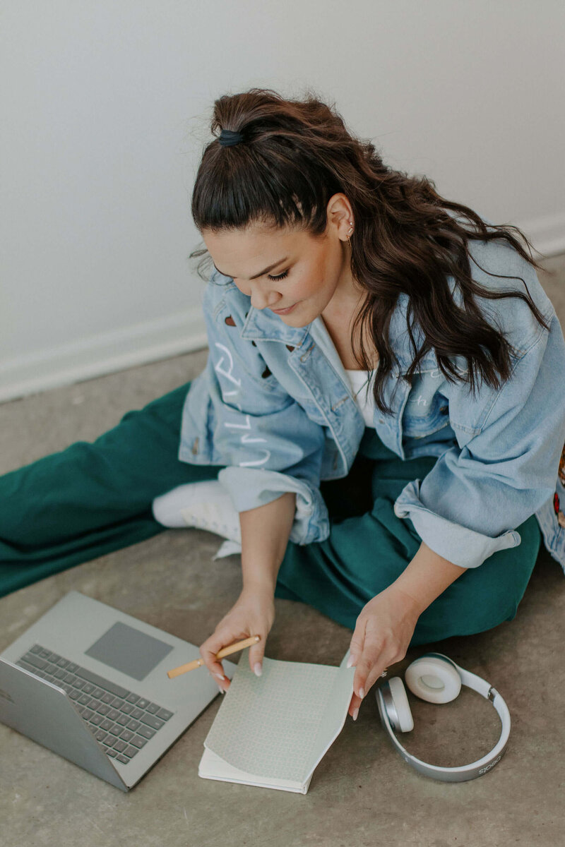 Woman with dark hiar in teal pants and jean jacket sitting on floor with laptop, headphones and journal