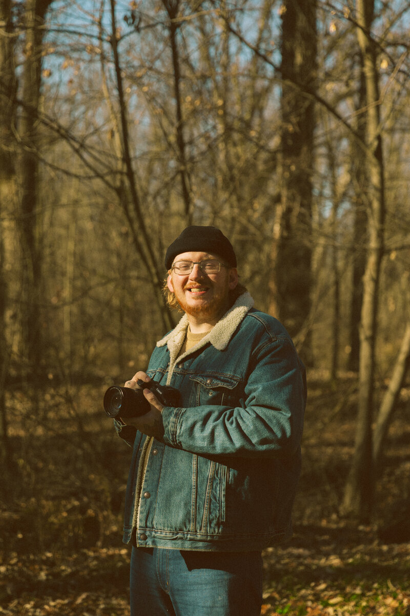a man smiles into the camera as he holds a camera in his tattoo covered hands