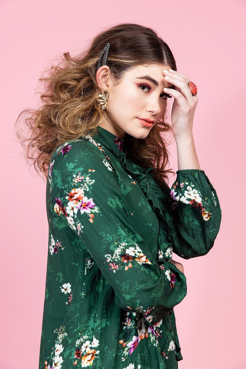 Teen model in a floral green blouse with voluminous curls and statement earrings posing against a pink studio backdrop – editorial-style image captured during a workshop