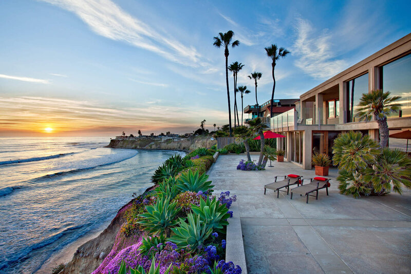A coastal backyard looking over a blue ocean in La Jolla with an orange sunset with patio furniture that has pops of red colors and palm trees in the background featured by Morganne Jones a San Diego Real Estate Agent in Point Loma