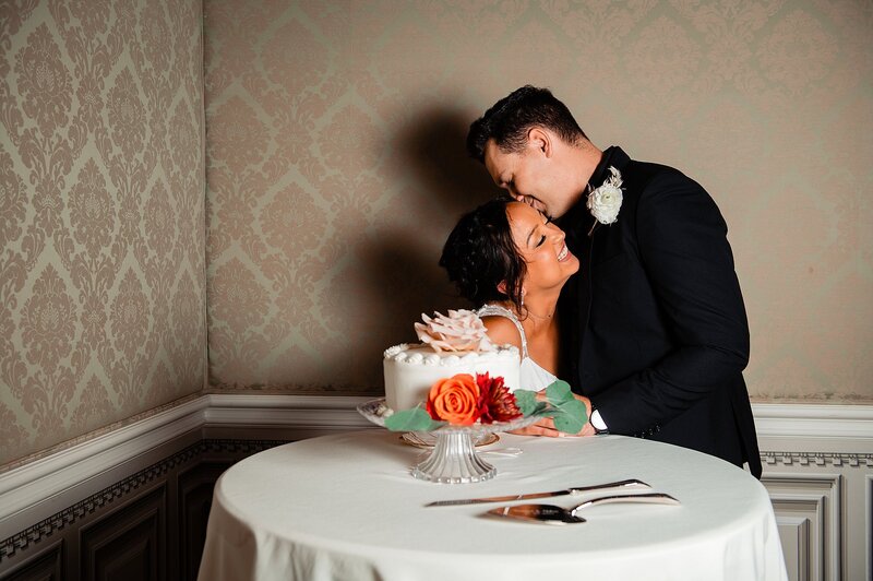 The bride and groom stand next to their wedding cake decorated with lush, orange and crimson roses. The groom, wearing a black suit with a white boutonniere leans in and whispers something into the bride's ear making her laugh at Oaklands Mansion.