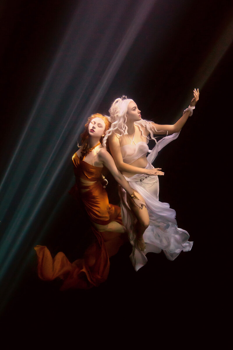 Two women gracefully pose underwater, illuminated by soft beams of light. One wears a flowing bronze gown, the other a sheer white wrap, creating an ethereal scene captured by Dallas Underwater Photographer Lindsay Walden.