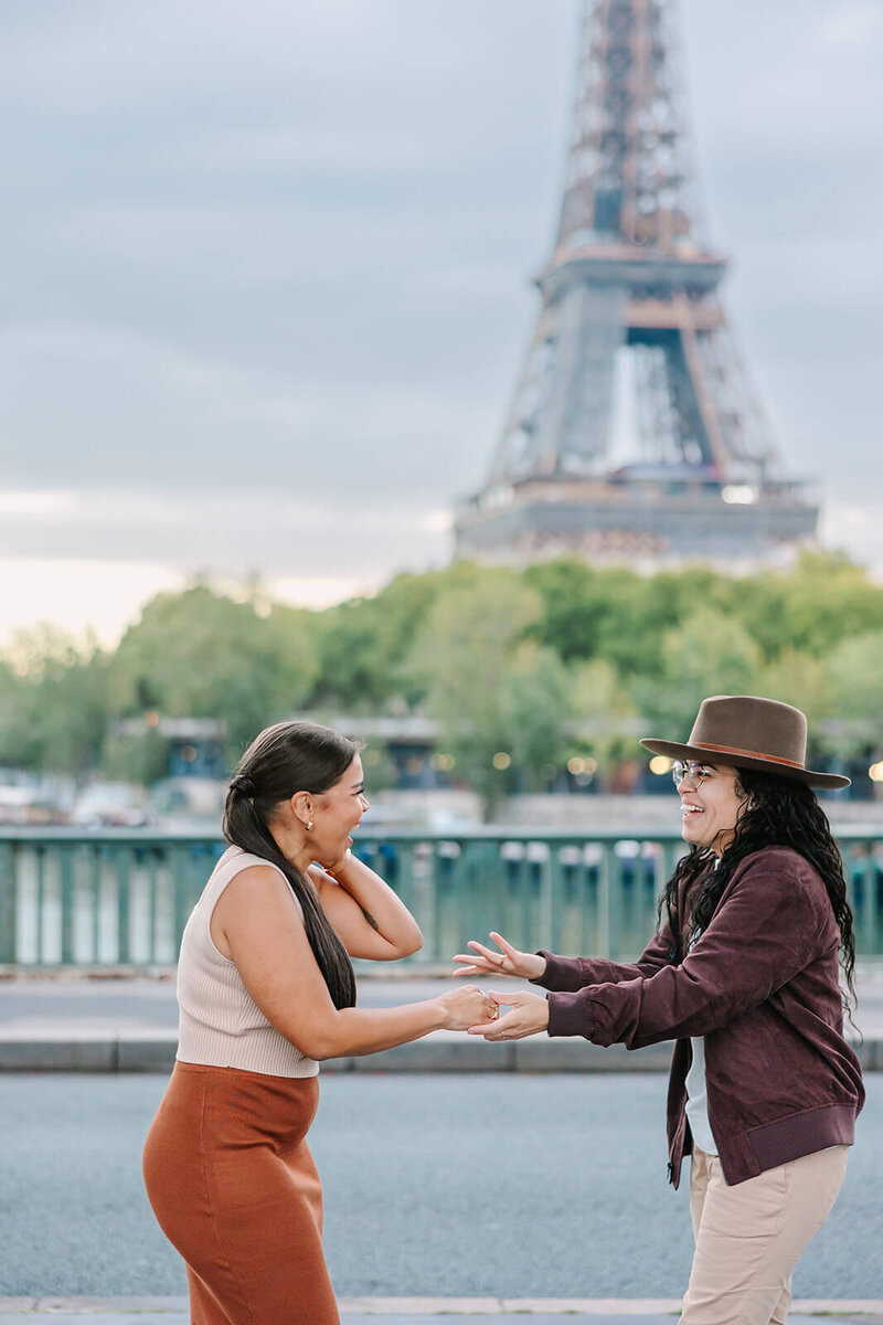 Excited lesbian couple holding hands after a proposal at the Eiffel Tower near Bir-Hakeim Bridge during a romantic Paris photoshoot.