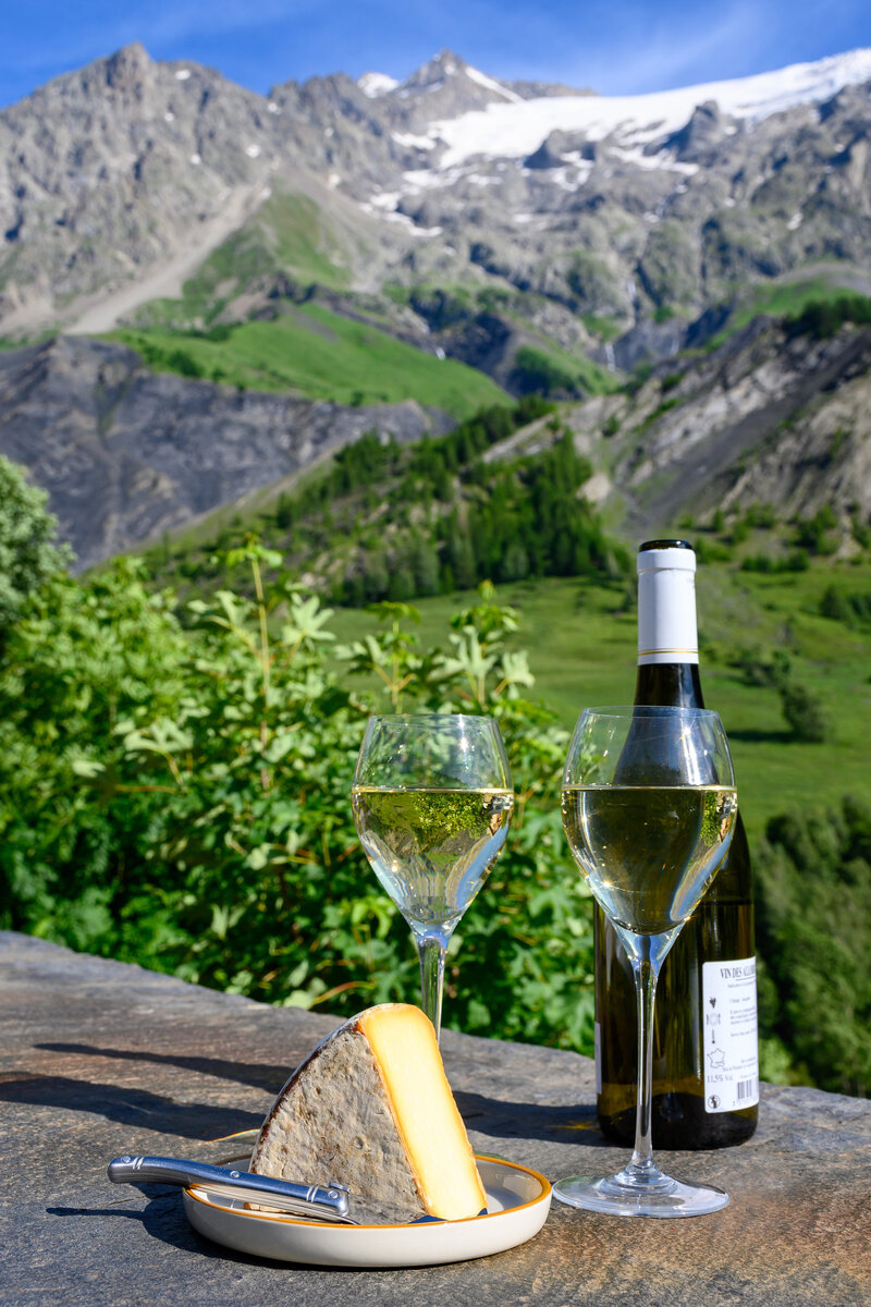 Cheese and wine, glasses of dry white Roussette de Savoie and Vin de Savoie wine from Savoy region with tomme cheese and view on Hautes Alpes mountains with snow on tops