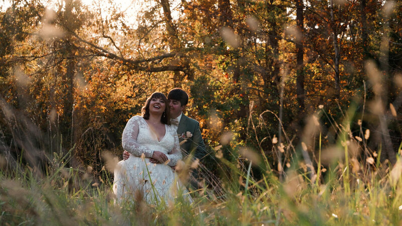 A newlywed couple with their arms around each other standing in tall grass 