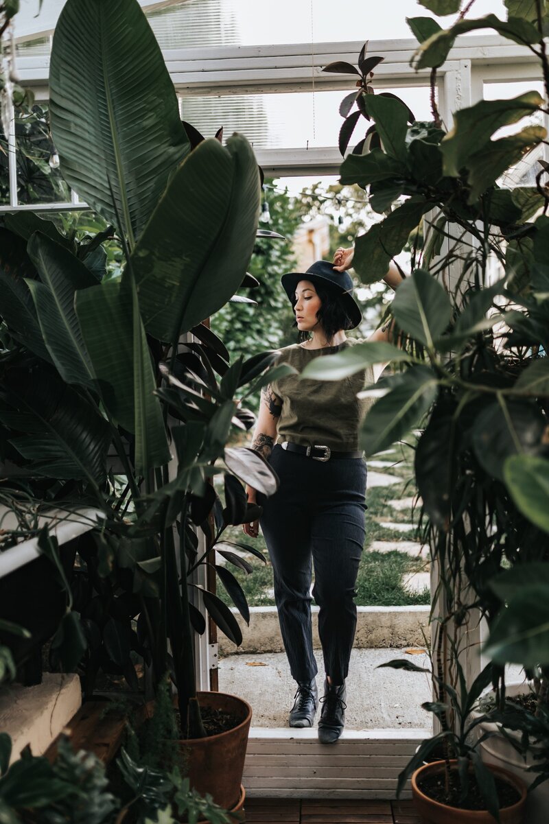 Woman in lush greenhouse wears a black hat, green top and black jeans while standing at the end of an aisle with an arm stretched above her.