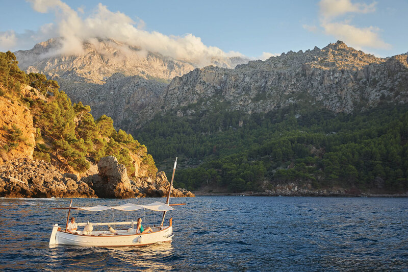 A small boat with a canopy floats on a calm sea near a rocky coastline. Pine trees cover the hills, and a mountain range with scattered clouds towers in the background under a clear sky.