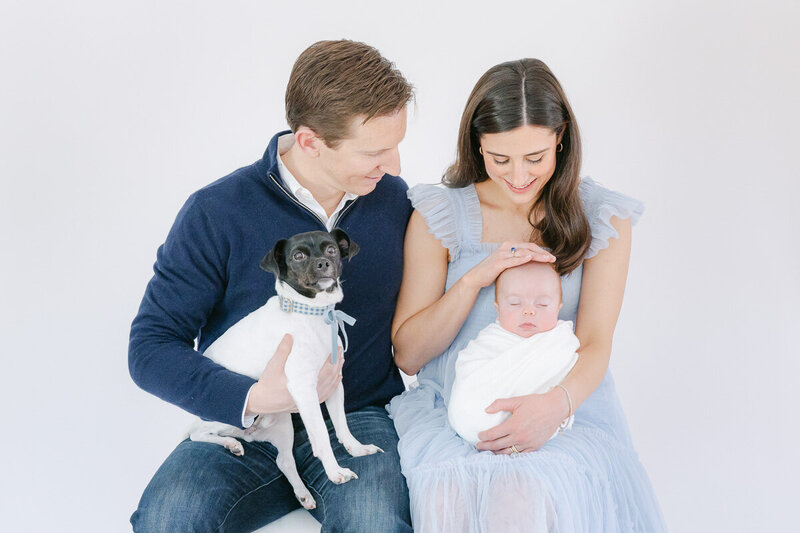A young couple poses lovingly with their newborn baby and small black-and-white dog during a bright, airy studio session. The mother, wearing a soft blue ruffled dress, cradles the swaddled baby while the father smiles holding their pup in his lap. Captured by Nicole Neira Photography, this image highlights the warmth and love of growing families—including their furry members.
