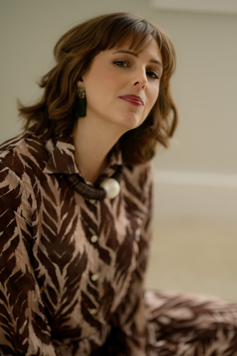 A woman with brown hair and bangs sits on the floor and leans toward the camera. She is wearing a brown and maroon shirt with leaves and dark, chunky jewelry.