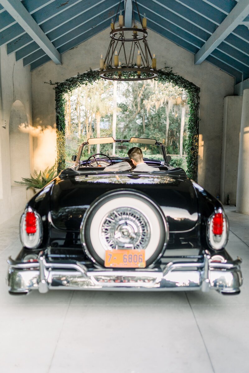 Bride rests her head on the grooms shoulder as they sit in a classic black car