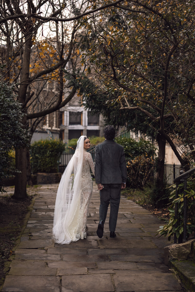 married couple take a moment of silence to walk hand in hand whilst the bridge looks back over her shoulder at the wedding photographer during their golden hour wedding shoot in central london