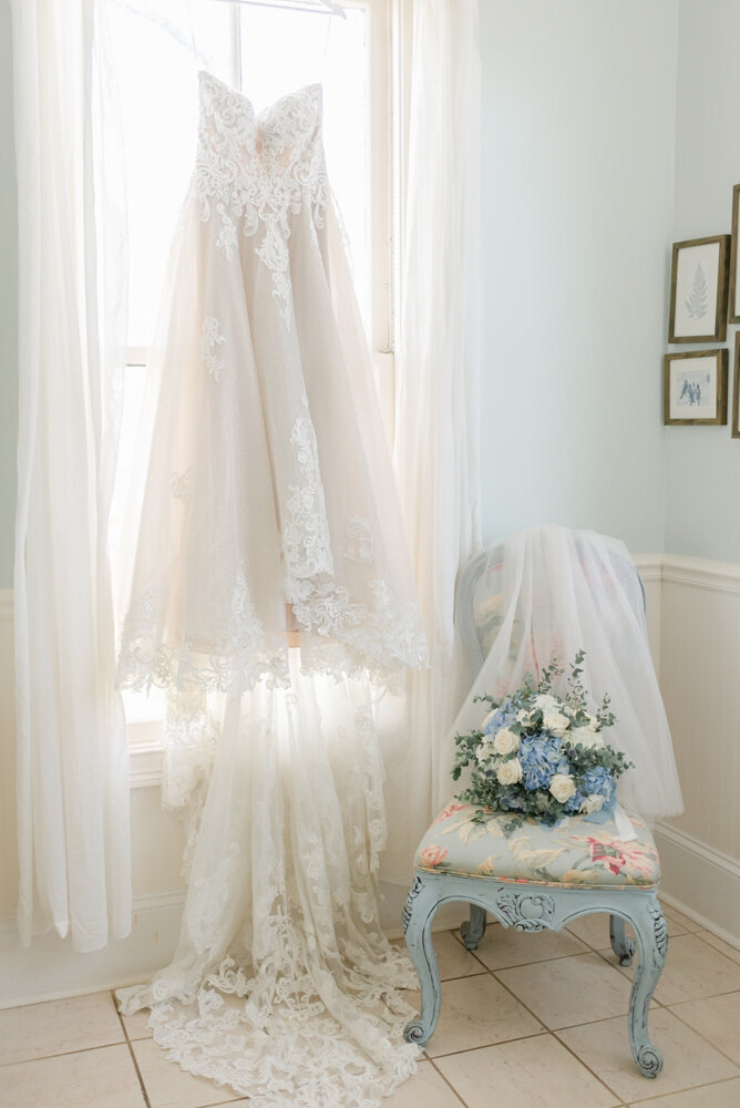 Lace wedding dress hanging in window with bouquet on chair next to it