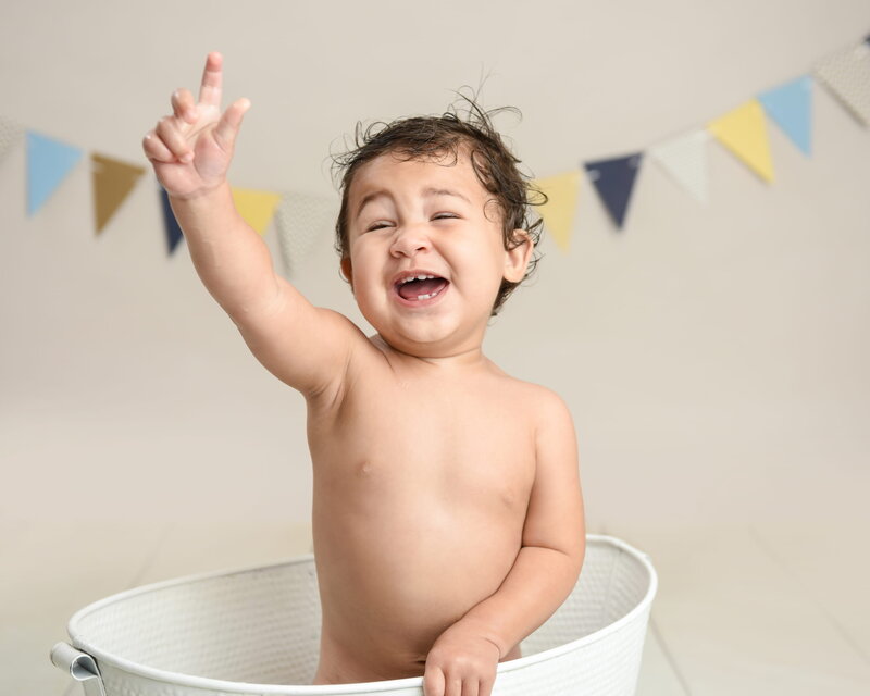 A cheerful baby laughing and pointing up while sitting in a white tub with yellow and blue bunting in the background.