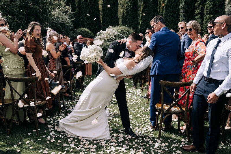 bride and groom do a dip kiss halfway down the aisle with white floral confetti at the winehouse queenstown