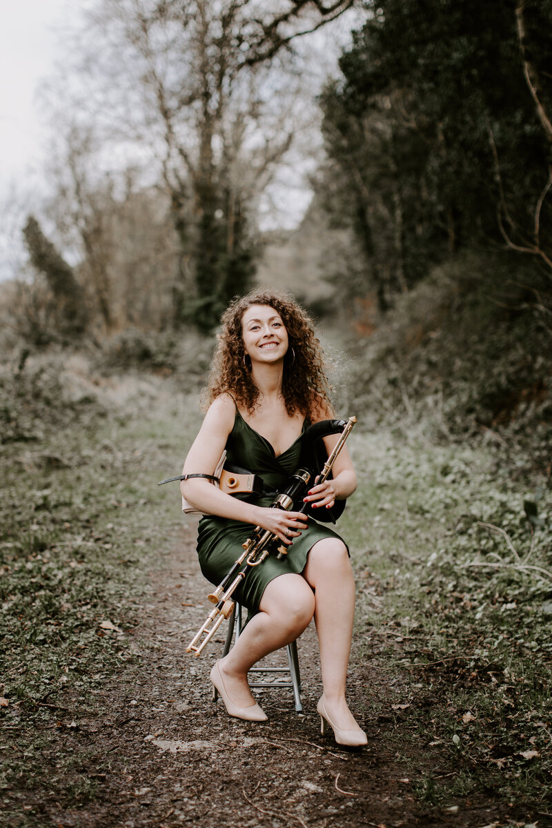 Seána Redmond smiling while seated on a woodland path, holding uilleann pipes, representing her traditional Irish music performances