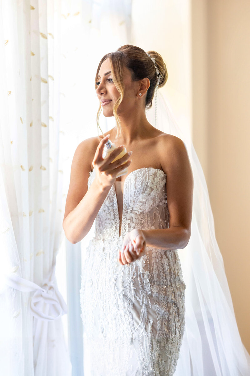 A bride in a detailed white wedding dress stands by a window, holding a perfume bottle. Her hair is elegantly styled, and she gazes outside with a serene expression. Sunlight softly illuminates the scene.