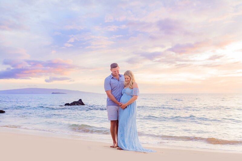 Pregnant woman in blue maternity dress standing on the shoreline on Maui and being kissed by her husband during their Maternity portraits in Wailea with Love + Water