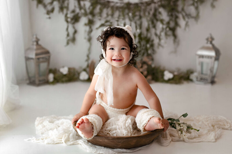 9 month  old toddler wearing  a bear bonnet  with matching  leg warmers  sitting  in a wooden  bowl