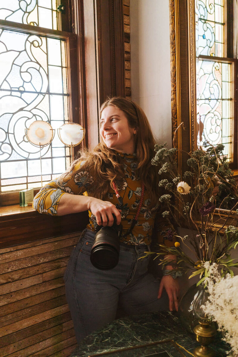 A person leaning against a windowsill smiling and holding a camera