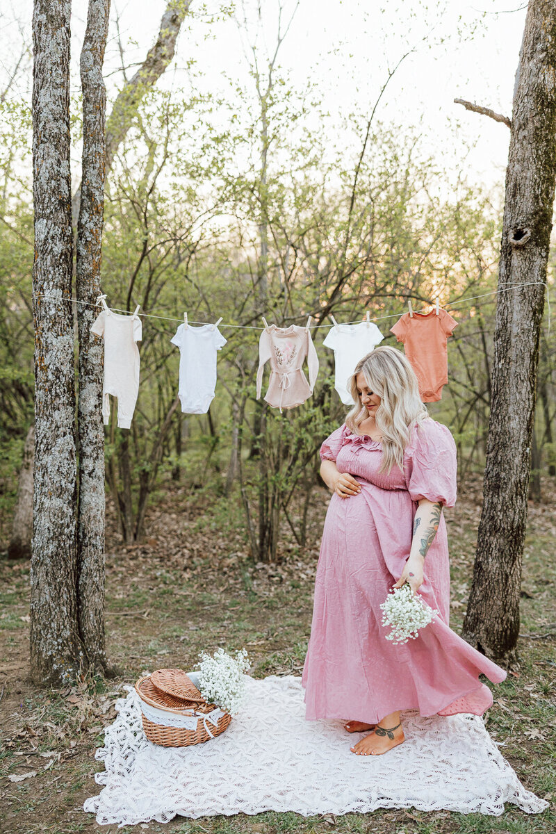 Themed maternity portrait of an expecting mother wearing a pink dress twirling while holding flowers with a clothes line, basket and newborn outfits in the background. 