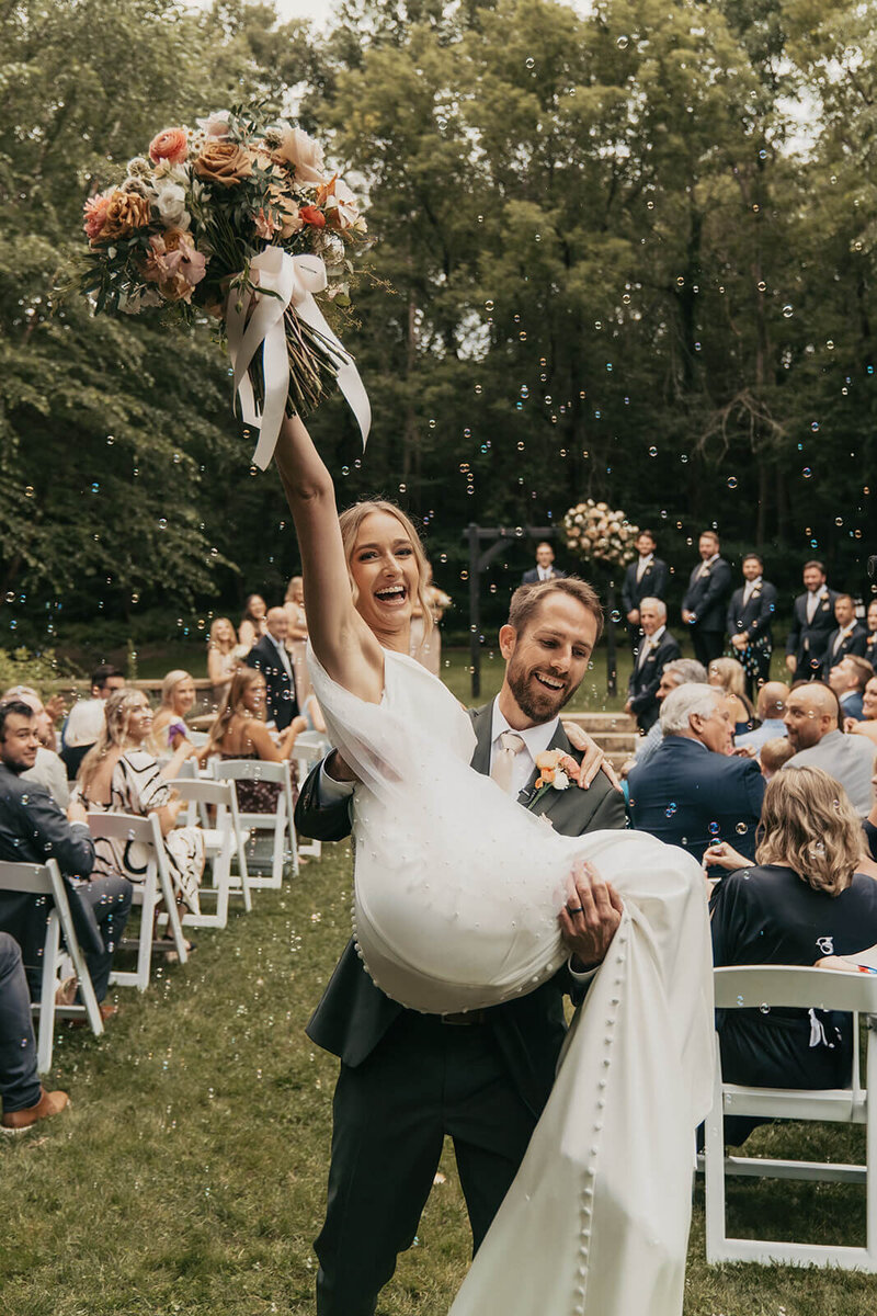 Bride raises her bouquet in the air as groom carries her down the aisle after they exchange their vows